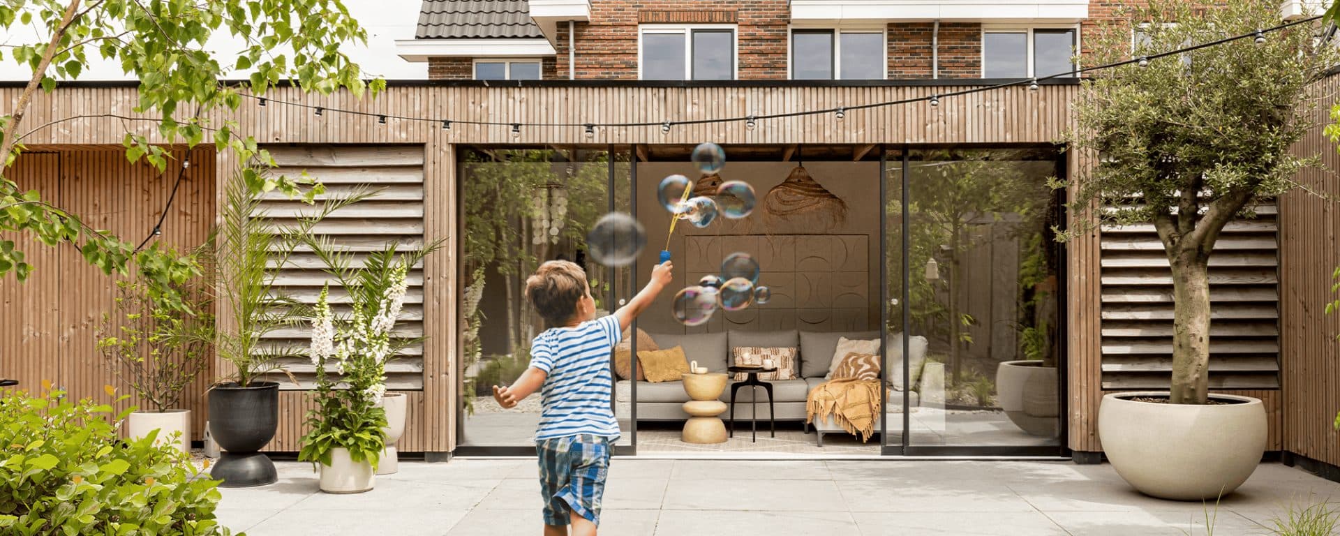 Kind blaast bellen voor een geopende glazen schuifwand in een houten gevel, met een zithoek erachter