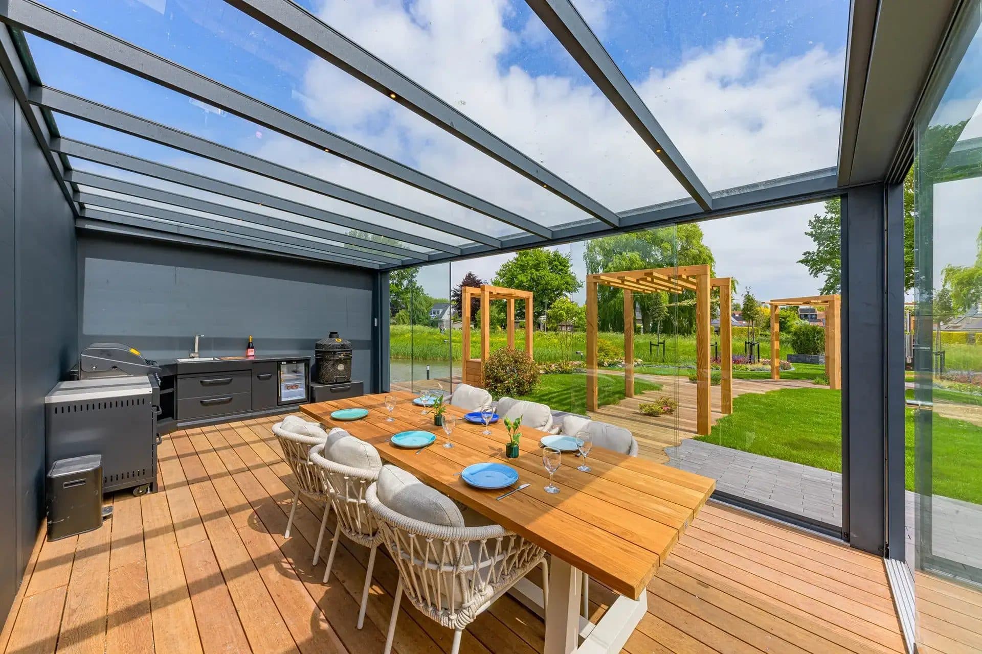 Gedekte eettafel en buitenkeuken onder een glasdak met glazen wanden, op een houten vlonder