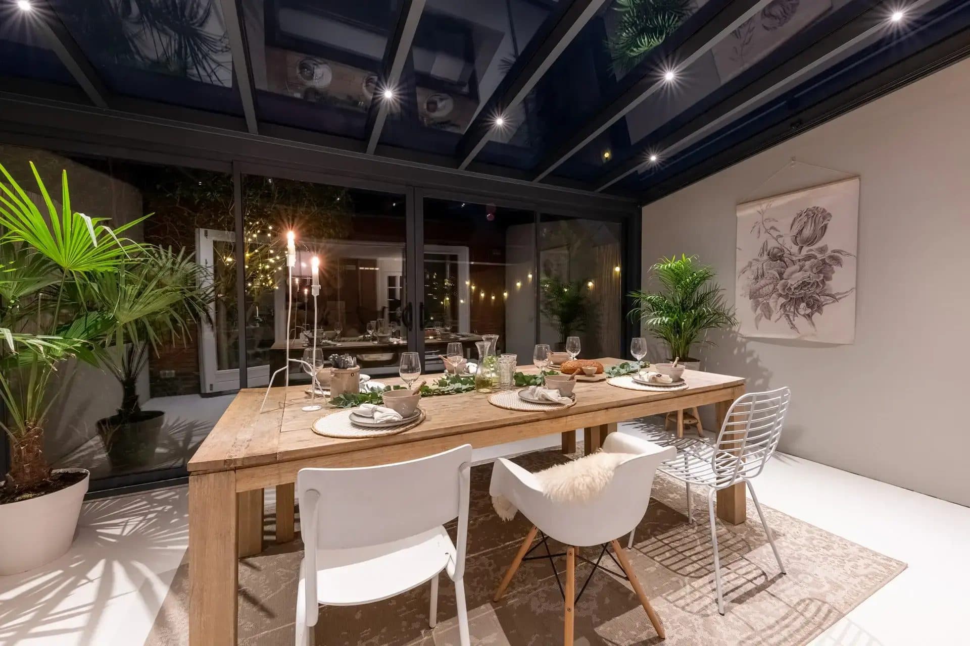 Gedekte houten eettafel met kaarsen in een tuinkamer met donker glasdak, ledspots en lichtslingers, 's avonds