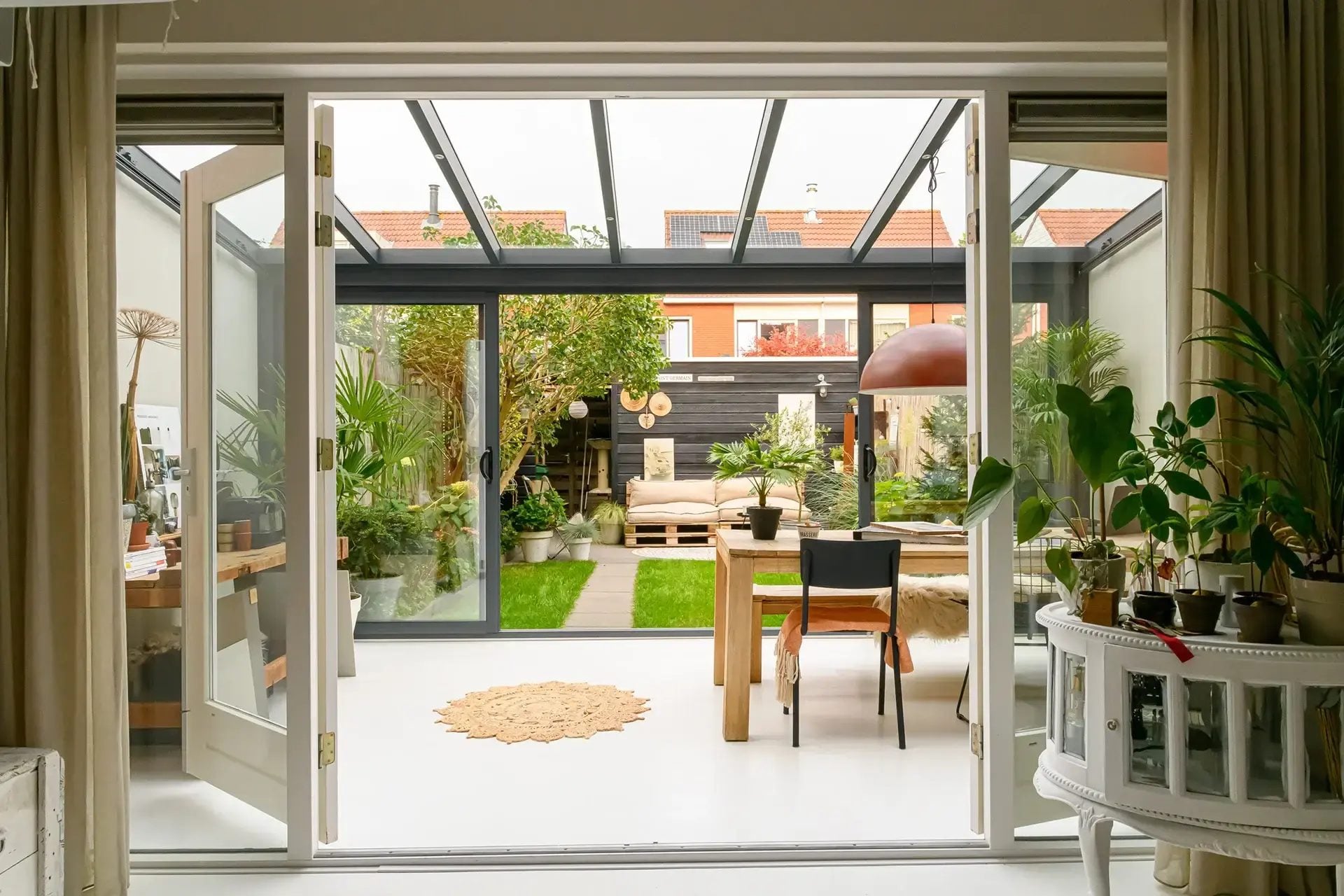 Zicht vanuit de woonkamer door openslaande deuren naar een tuinkamer met glasdak, eettafel en planten, en daarachter de tuin