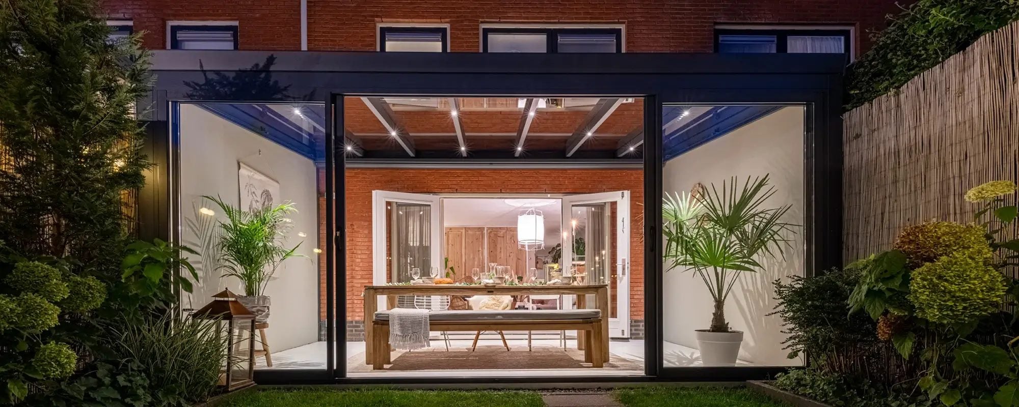 Tuinkamer met glazen wanden bij schemering, met brandende spots in het dak boven een gedekte eettafel en planten