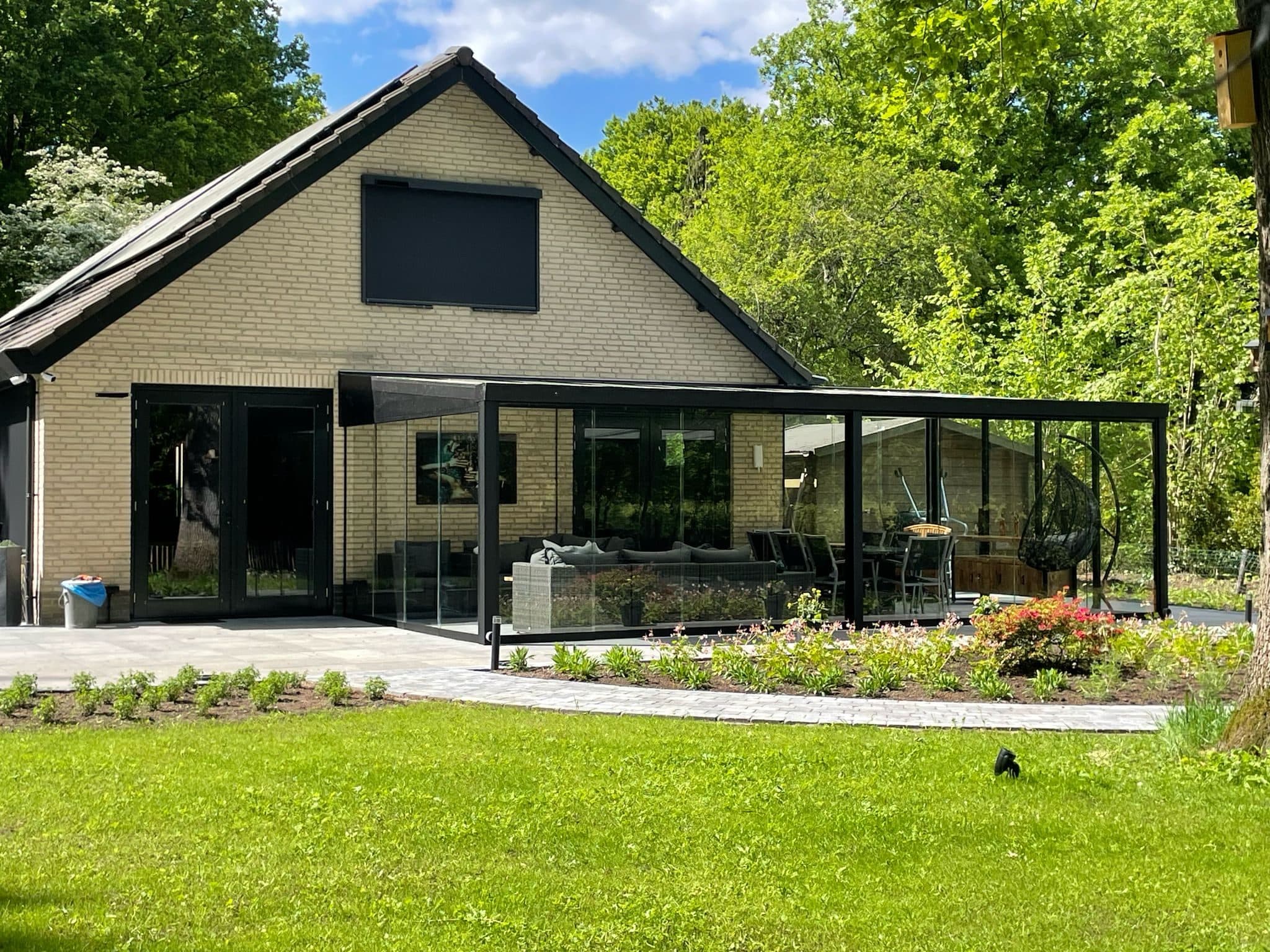 Zwarte veranda met glazen schuifwanden langs de achtergevel van een woning met lichte baksteen, met een loungeset, een hangstoel en een nieuwe border ervoor