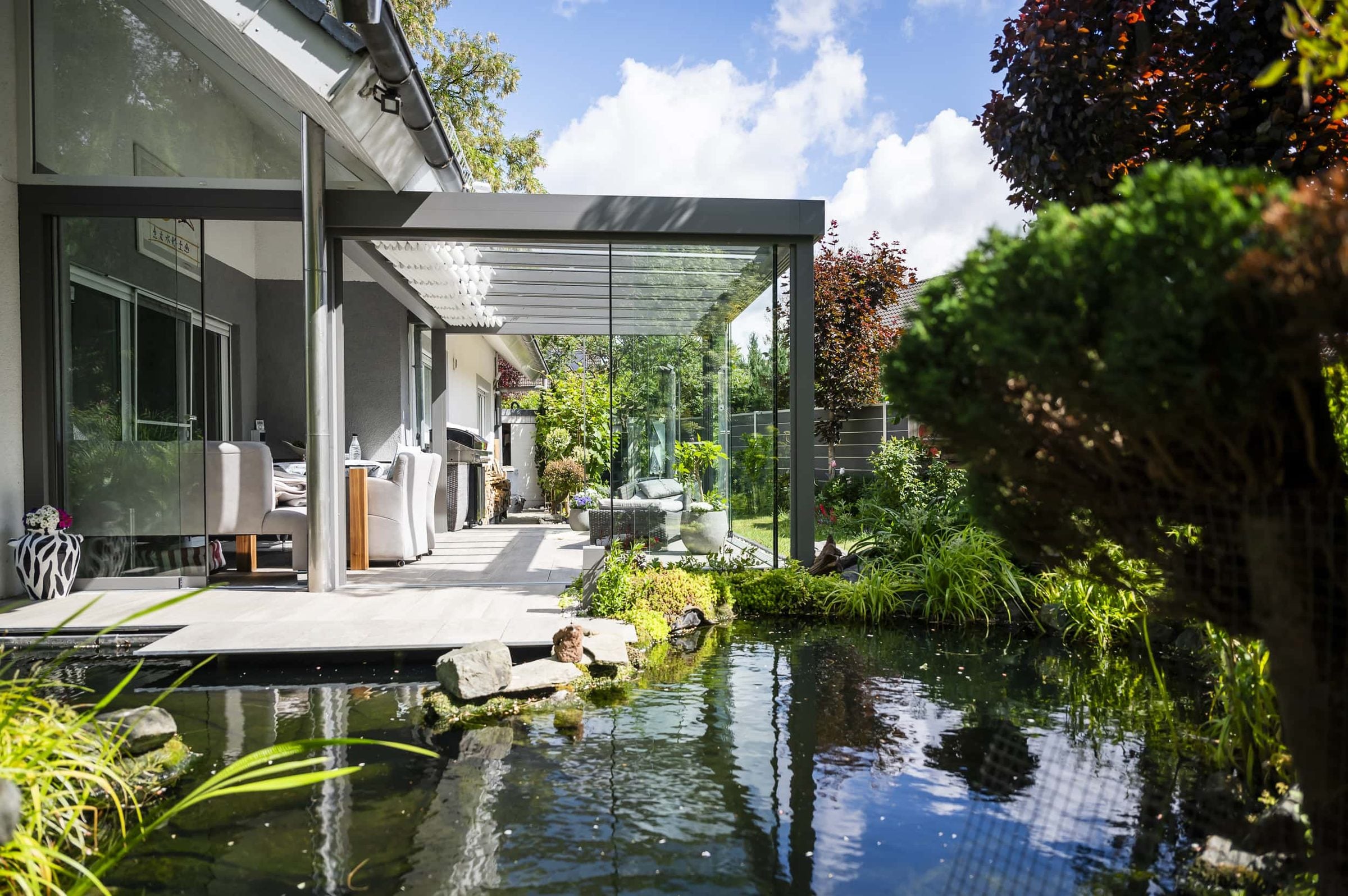 Antraciet veranda met strakke goot en glazen wanden langs een tuinvijver met waterplanten