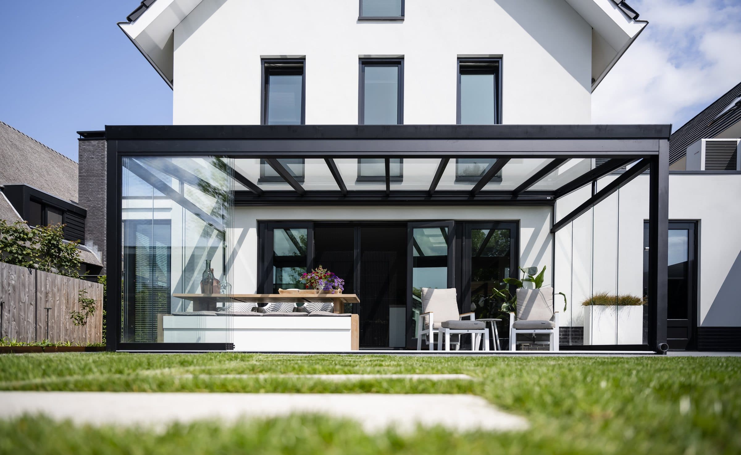 Zwarte veranda met glasdak en glazen wanden tegen een witte woning, gezien vanaf het gazon