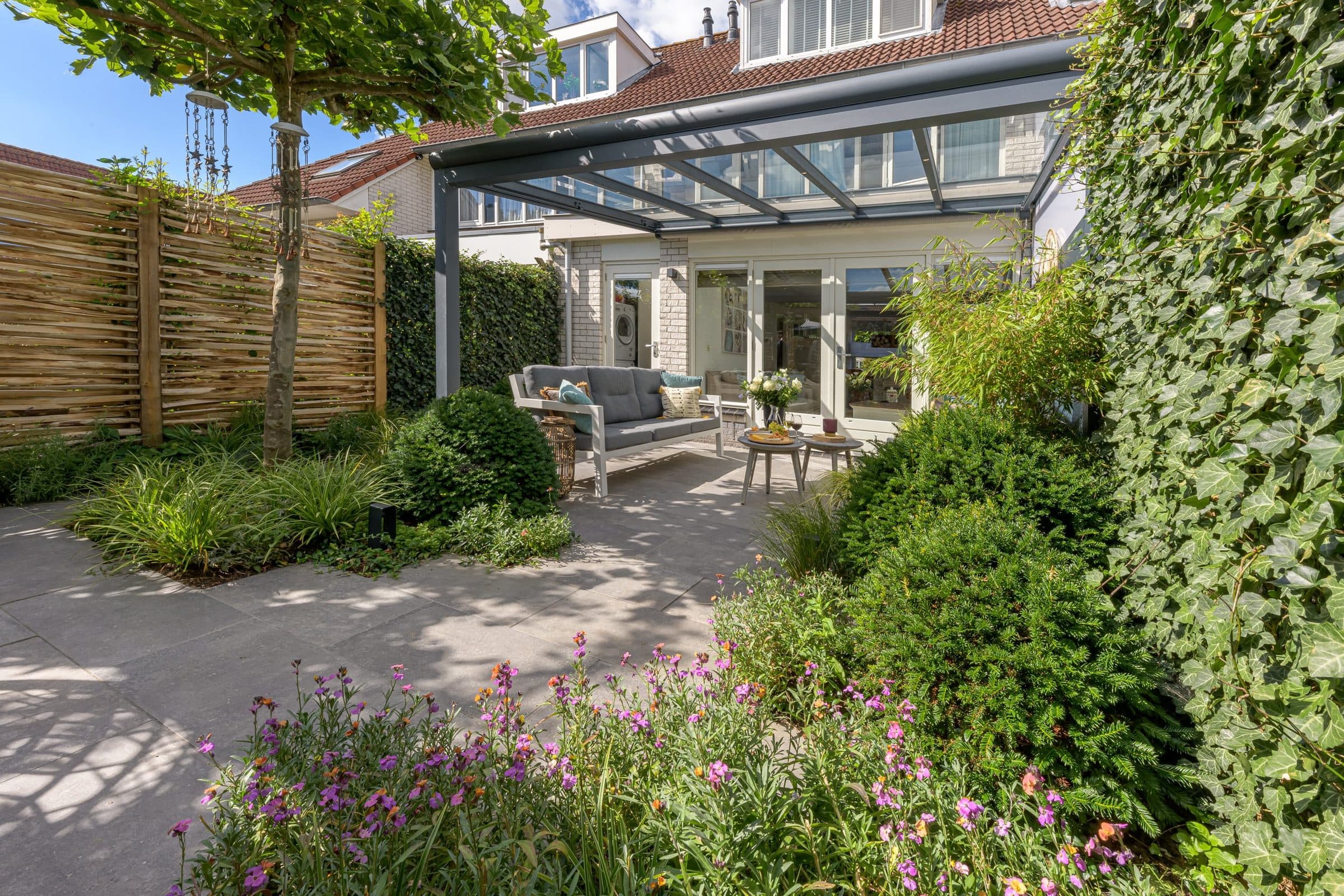 Antraciet veranda met glasdak achter een woning, gezien door een weelderige tuin met klimop en bloeiende planten