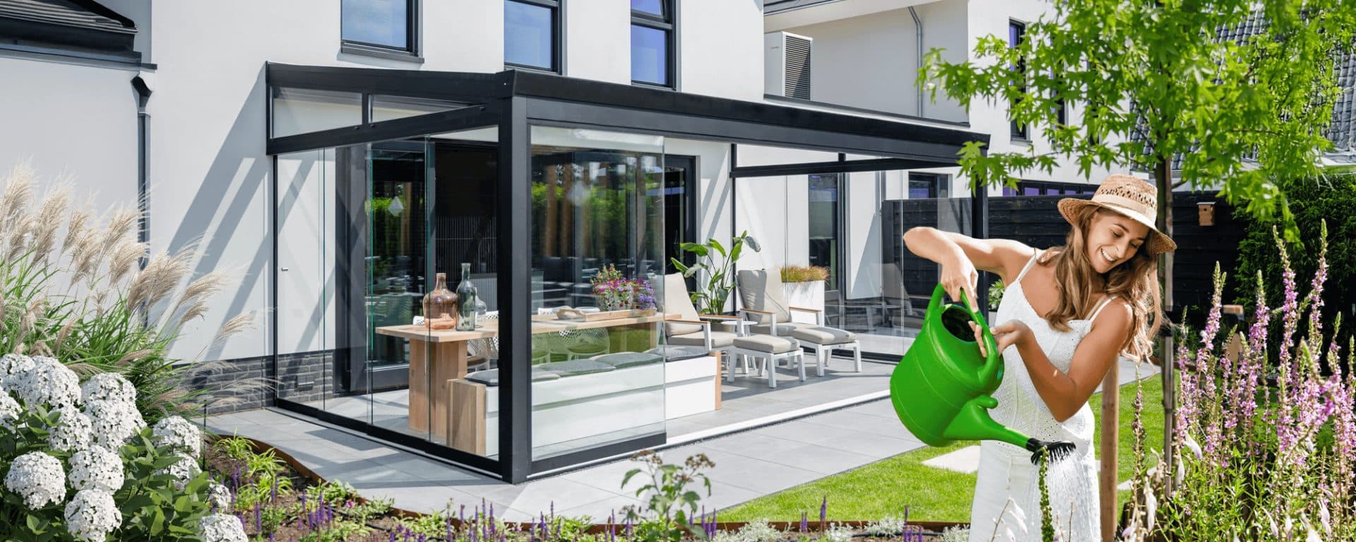 Vrouw geeft planten water naast een antraciet overkapping met glazen wanden aan een witte woning