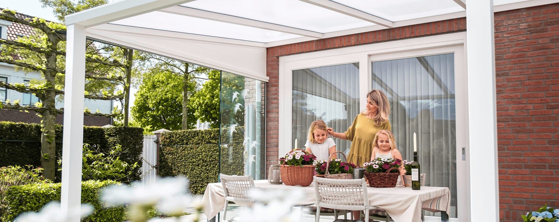 Moeder en twee dochters zetten bloemen op een gedekte tafel onder een witte veranda met glazen wanden