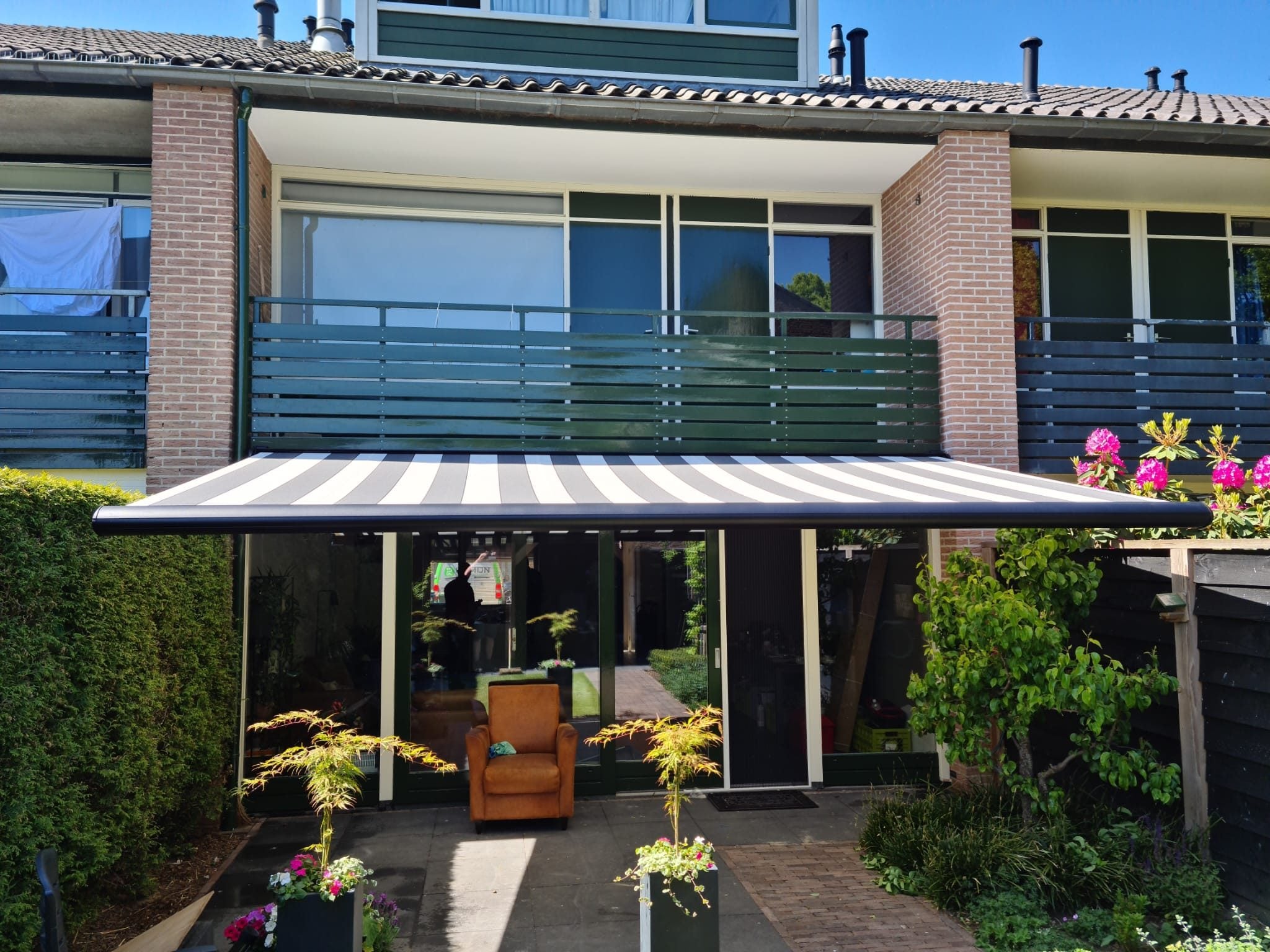 Uitgerold knikarmscherm met grijs-wit gestreept doek boven het terras van een rijwoning met balkon
