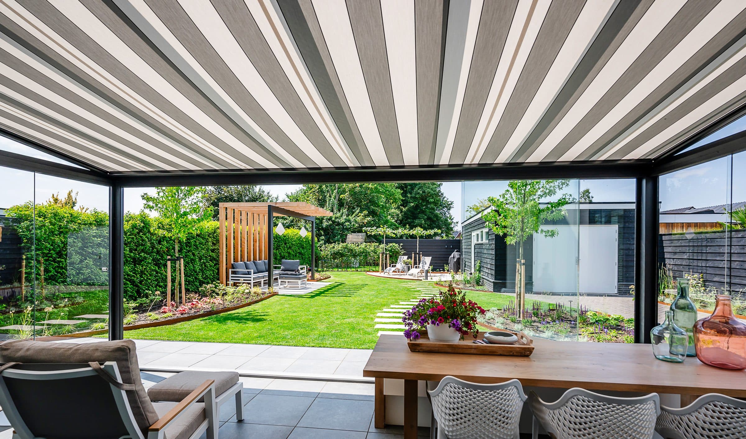 Grijs-wit gestreepte onderdakzonwering boven een eettafel en loungestoel, met zicht op een aangelegde tuin